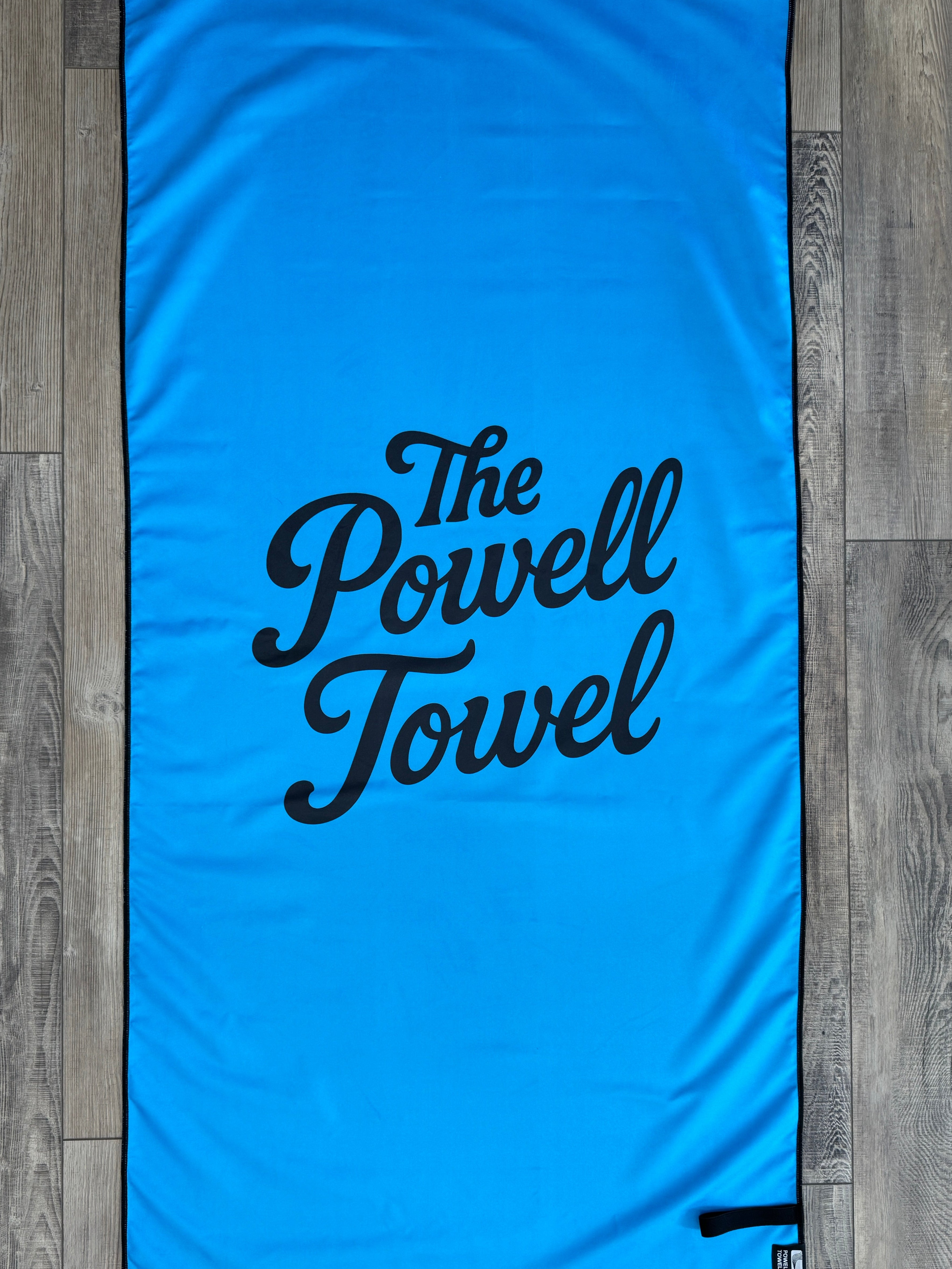Blue towel with 'The Powell Towel' text on a wooden floor